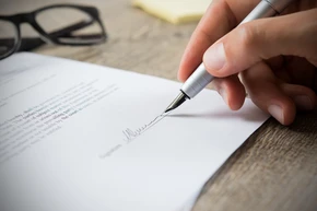 Une main tient un stylo plume et signe un document sur un bureau en bois
