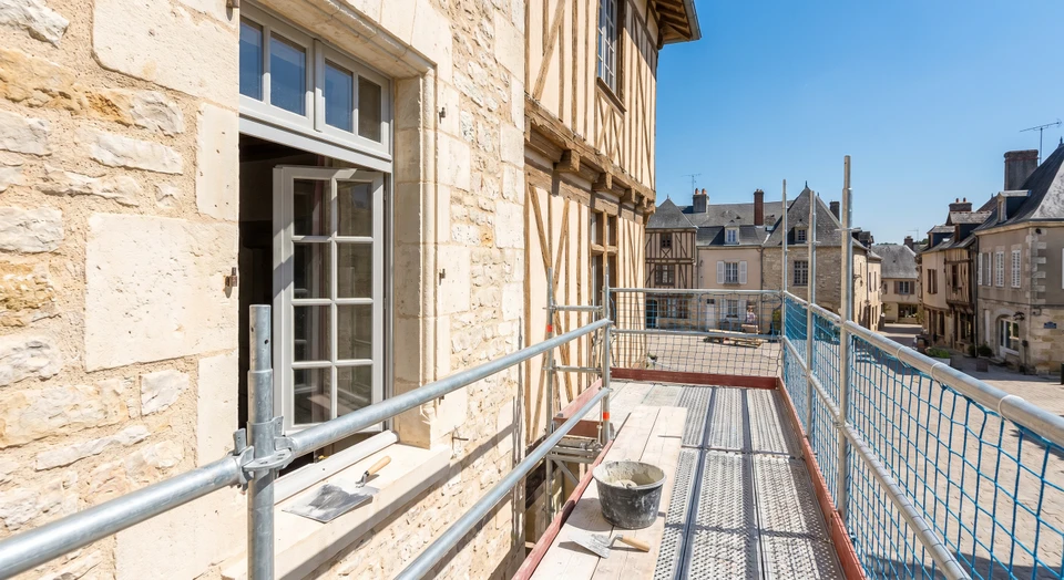 Vue d'un chantier de rénovation intérieure en cours, montrant plusieurs corps de métiers interagissant ou ayant travaillé dans un même espace : un ouvrier maçon/démolition en arrière-plan, des matériaux de construction et outils dispersés, et un carreleur ou peintre au premier plan. L'ambiance doit montrer une coordination professionnelle et organisée avec plusieurs étapes du projet visibles simultanément, donnant l'impression d'une gestion cohérente et intégrée du chantier.